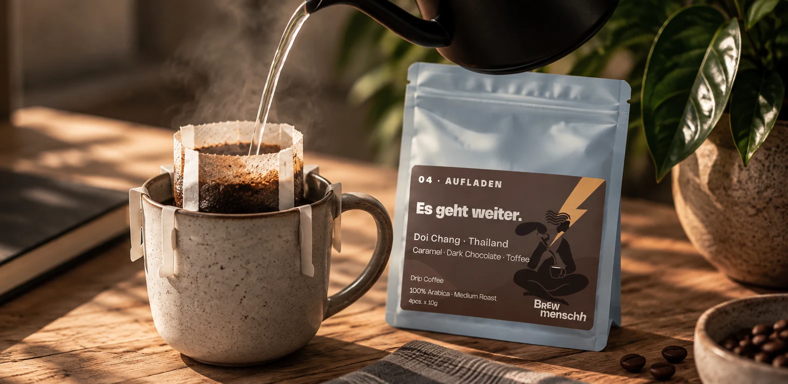 Water poured into a drip bag on a ceramic mug beside a BrewMensch Aufladen package, a book and a plant on a sunlit wooden table